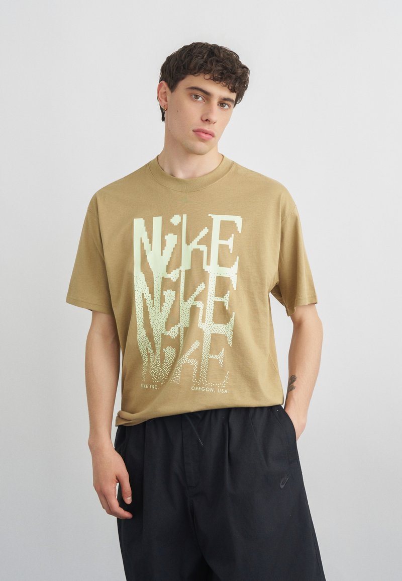 Νέος άνδρας με κοντά σγουρά μαλλιά, φοράει μια μπεζ μπλούζα Nike με μεγάλο λευκό γραφικό κείμενο και μαύρα σορτς Nike, στέκεται μπροστά σε απλό φόντο.