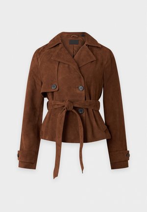 Une femme porte une veste marron ceinturée, un pantalon en cuir noir, des chaussures marron et un sac à bandoulière sur un fond de studio uni.