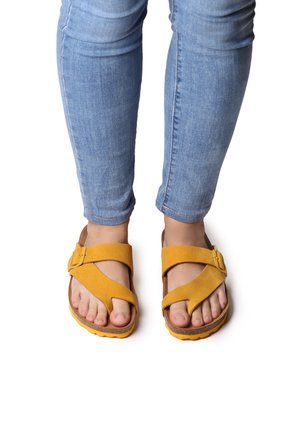 Sandali in suede giallo con design a strap incrociati, accentati da marrone e suola texture, visti frontalmente con jeans di denim usurati.