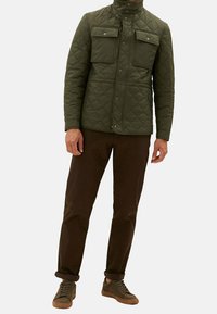 Veste matelassée vert olive avec deux poches poitrine, poches latérales zippées, fermeture à pression et col montant ; associée à un pantalon marron et des baskets marron.
