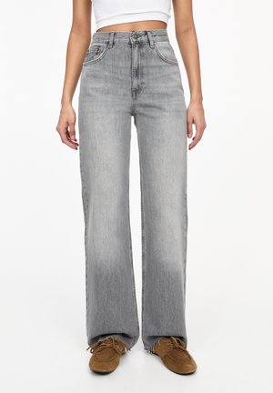 Frau trägt hoch taillierte, weit geschnittene graue Jeans, weißes, bauchfreies Tanktop und braune Schnürschuhe, steht vor einem schlichten weißen Hintergrund.