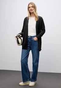 Svart kofta över en vit topp, kombinerad med vida blå jeans. Modellen håller en randig beige väska. Mjukt tyg och avslappnad stil.