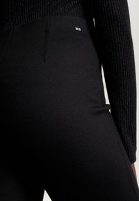 Leggings noirs en tissu extensible avec une texture lisse, présentant un petit détail de logo près de la taille. Le design est ajusté.