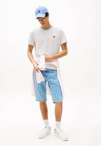 Ung mand iført lyseblå cap, stribet t-shirt, lyseblå denimshorts, hvide sneakers og en pink skjorte bundet omkring livet, stående foran en hvid baggrund.