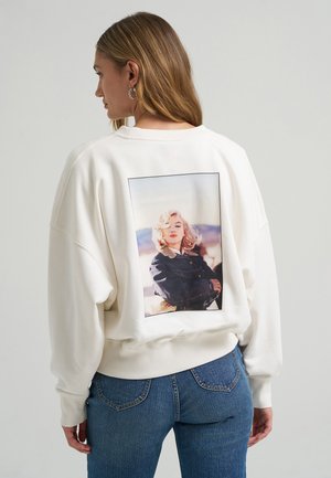 Žena s dlhými blond vlasmi nosí bielu mikinu s vintage fotografiou blond ženy v čiernej bunde potlačenou na chrbte.
