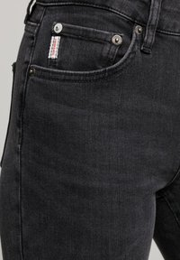 Los jeans de mezclilla negros cuentan con un diseño clásico de cinco bolsillos, remaches plateados y una pequeña etiqueta roja en el bolsillo frontal. Tienen una textura suave y un corte ajustado.