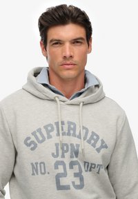 Man met donker haar in een lichtgrijze hoodie met de tekst "SUPERDRY JPN NO. 23 OPT" over een blauwe overhemd tegen een witte achtergrond.