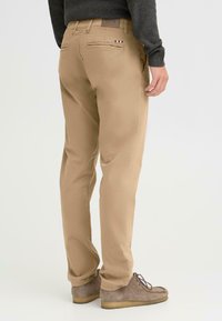 Khaki chinos dengan potongan lurus, saku depan dan belakang, serta aksen logo merek yang halus. Kain terlihat halus dengan finishing yang rapi.