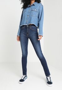 Chaqueta de mezclilla recortada de color azul claro con dos bolsillos, combinada con jeans ajustados oscuros. Zapatillas blancas con cordones y acentos en navy.