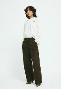 Long-sleeve white shirt with circular logo, paired with dark green wide-leg corduroy pants and green heeled ankle boots. Plain background.