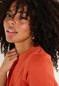 Suéter de punto naranja con cuello redondo, tejido texturizado, ligera canalé en el cuello y rizos sueltos enmarcando la cara. Arete dorado en forma de aro visible.