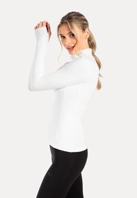 Weißes langärmliges Sportoberteil mit hohem Kragen und körperbetontem Schnitt. Weiche Textur und Reißverschluss-Detail an der Vorderseite, kombiniert mit schwarzen Leggings.