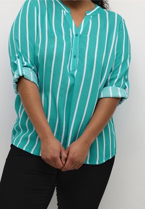 Persona con una camisa de rayas verticales en color verde azulado y blanco, con las mangas remangadas, de pie con las manos entrelazadas al frente.