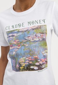 Vit bomull t-shirt med en färgglad tryck av Monets "Vattenliljor" med grön text ovanför och nedanför konstverket.