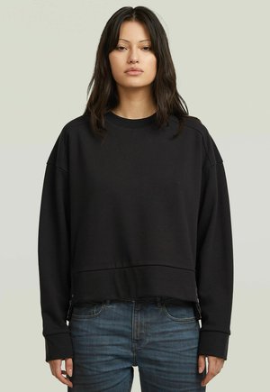 Vrouw met lang donker haar, gekleed in een effen zwarte oversized sweatshirt en blauwe jeans, staand tegen een neutrale achtergrond.