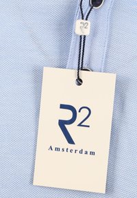 Lichtblauw poloshirt met een textuurstof, twee donkere knopen en een label met "R2 Amsterdam" in marineblauwe letters.