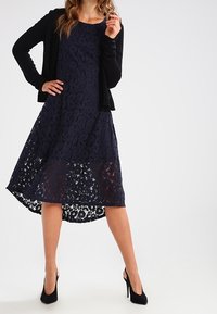 Krajkové šaty v námořnické modři s vysokým předním a nižším zadním lemem, doplněné černým kardiganem. Vyznačují se květinovými vzory a volným střihem. Nositelná s černými lodičkami.