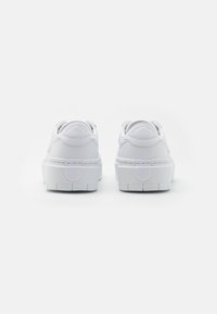 Baskets blanches avec une finition en cuir lisse, bout arrondi et semelle épaisse et texturée. Présente un détail de logo subtil sur le passant de talon.