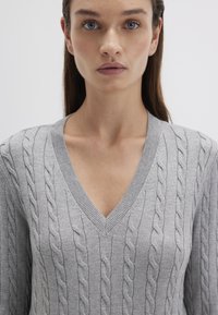 Helles grauer Kabelstrickpullover mit tiefem V-Ausschnitt, ausgestattet mit gerippten Bündchen und Saum. Das strukturierte Muster verleiht Tiefe und visuelles Interesse.