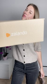 Zalando márkájú kartondoboz, amelyet elöl tartanak; a doboz világosbarna, és rajta látható a logó. A személy szürke kötött felsőt és sötét farmernadrágot visel.