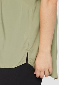 Olivegrüne Bluse mit lockerer Passform, kurzen Ärmeln, seitlichen Schlitzen und glatter Textur, kombiniert mit schwarzen, strukturierten Leggings.