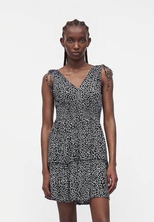 Γυναίκα με πλεξούδες, που φοράει ένα μαύρο αμάνικο φόρεμα με λευκό λουλουδάτο μοτίβο και δεσίματα στους ιμάντες, στέκεται μπροστά από ένα απλό λευκό φόντο.