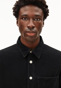 Zwarte corduroy shirt met een knoopsluiting aan de voorkant, kraag en een borstzak. Heeft textuur met verticale lijnen en witte knopen.