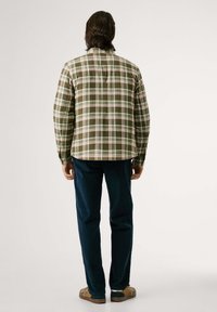 Camisa de franela a cuadros verdes con detalles en blanco y rojo, con puños abotonados y un dobladillo curvo, combinada con pantalones oscuros azules y sandalias marrones.