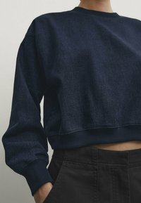 Laivastonsininen, denimiä oleva lyhyt collegepaita. Siinä on pyöreä pääntie, resorit hihansuissa ja helmassa, sileä pinta ja hillitty kiilto.