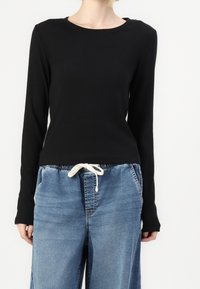 Schwarzes Langarmshirt mit rundem Halsausschnitt, aus glattem Stoff; kombiniert mit lockeren blauen Jeanshosen mit elastischem Bund und Kordelzug.