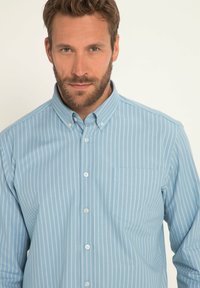 Jasnoniebieska koszula button-up w białe pionowe paski, z kołnierzykiem i kieszonką na lewej piersi. Gładka faktura materiału.