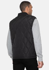 Gilet trapuntato nero con colletto alto, caratterizzato da una cucitura a motivo a diamante. Indossato sopra una maglietta a maniche lunghe grigia, con polsini elasticizzati.