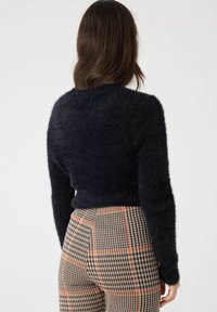 Mujer con cabello castaño que lleva un suéter negro esponjoso y pantalones de cuadros beige con acentos naranjas, de espaldas contra un fondo simple.