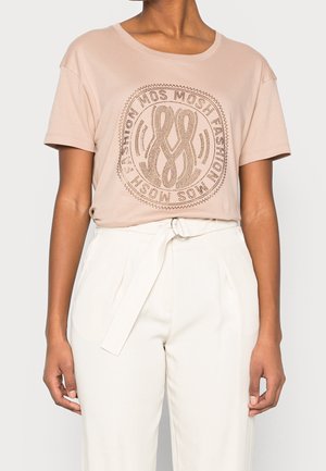 Vrouw draagt een lichtroze T-shirt met korte mouwen en een MOS MOSH bedrukt ontwerp, gecombineerd met witte, hooggetailleerde broek met een geknoopt stoffen ceintuur.