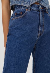 Džíny z modré denimu s vysokým pasem, rovný střih nohavic, viditelným švem, dvěma předními kapsami a kovovou stahovací knoflíkovou zapínací. 