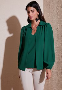 Blusa verde con cuello de camisa, mangas abullonadas y un diseño de escote en V. El tejido parece suave y ligero. Usada sobre pantalones de color claro.