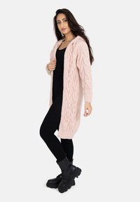 Cardigan en tricot rose clair avec un motif en câble, des manches longues et une capuche, superposé à un haut noir ajusté et un legging noir.