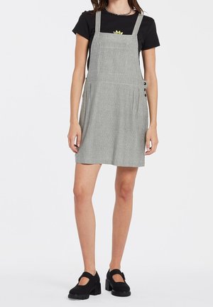 Femme portant un t-shirt noir sous une robe salopette gris à fines rayures avec des détails à boutons, associée à des chaussures Mary Jane noires.