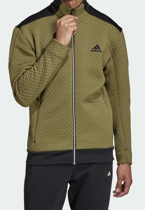 Mężczyzna ubrany w oliwkową, fakturowaną kurtkę rozpinaną z czarnymi ramionami i logo Adidas, połączoną z czarnymi spodniami Adidas.