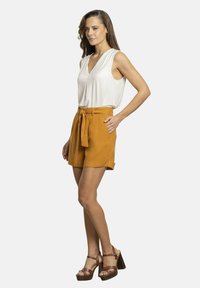 Top blanco sin mangas con escote en V, combinado con pantalones cortos naranjas de cintura alta que cuentan con un cinturón de lazo. Zapatos altos marrones con tiras. Diseño minimalista.