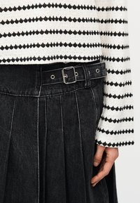 Μαύρη πλισέ φούστα με ζώνη και ασημί αγκράφα, φορεμένη με λευκό πουλόβερ που έχει μαύρο διαμαντένιο σχέδιο.