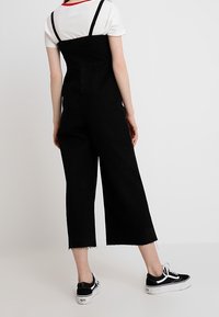 Svart denim jumpsuit med vida ben och axelremmar, med fransad nederkant och bärs med svarta sneakers. Enkel, avslappnad design.