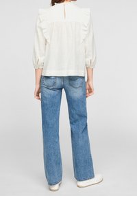 Blusa bianca a balze con dettagli a chiave sulla schiena, abbinata a jeans azzurri a vita alta. Vestibilità casual con cuciture visibili sui jeans.
