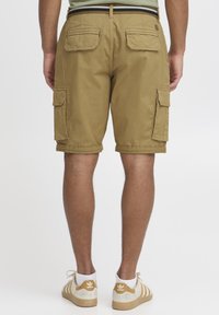 Shorts cargo kaki avec plusieurs poches latérales, revers roulés et un tissu texturé ; associés à des baskets blanches avec des accents dorés.
