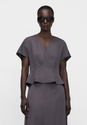Frau trägt übergroße schwarze Sonnenbrille und ein enganliegendes graues Kleid mit kurzen Ärmeln und einem Peplum-Taillenbund, steht vor einem schlichten Hintergrund.
