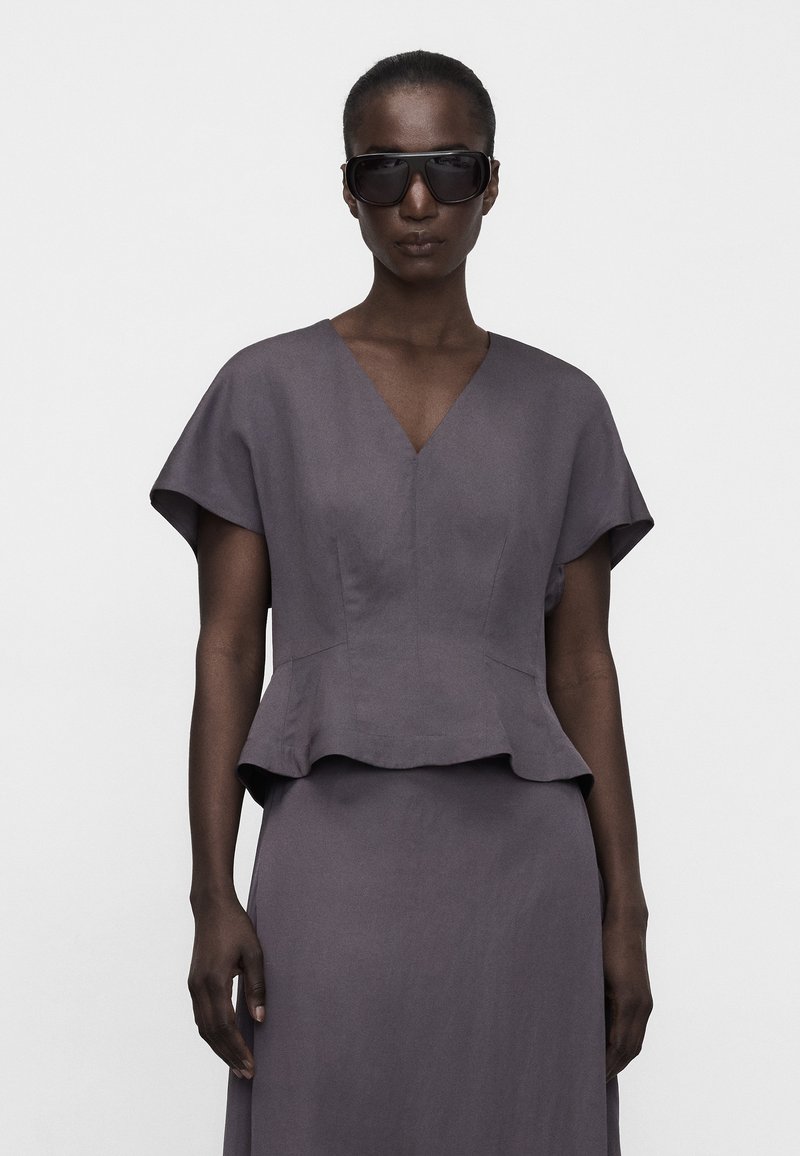 Woman wearing oversized black sunglasses and a fitted gray dress with short sleeves and a peplum waist, standing against a plain background.