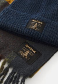 Navyblauwe gebreide muts en donkergrijze wollen sjaal met franjes, beide met een zwart Barbour-logo met een ontwerp van een vuurtoren.