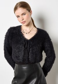 Jonge vrouw met een lichte huid en blauwe ogen, gekleed in een zwarte fluffy cropped trui, een zwarte leren rok en een gouden charmeketting.
