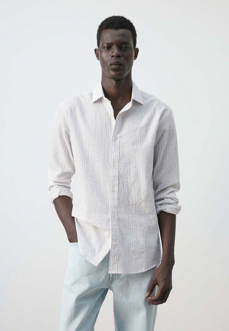 Jeune homme à la peau foncée et aux cheveux courts portant une chemise blanche texturée à boutons et un jean bleu clair, une main dans la poche.