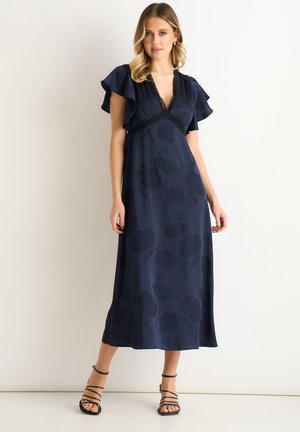 Woman standing against a white background wearing a navy blue midi dress with flutter sleeves and black strappy heels.